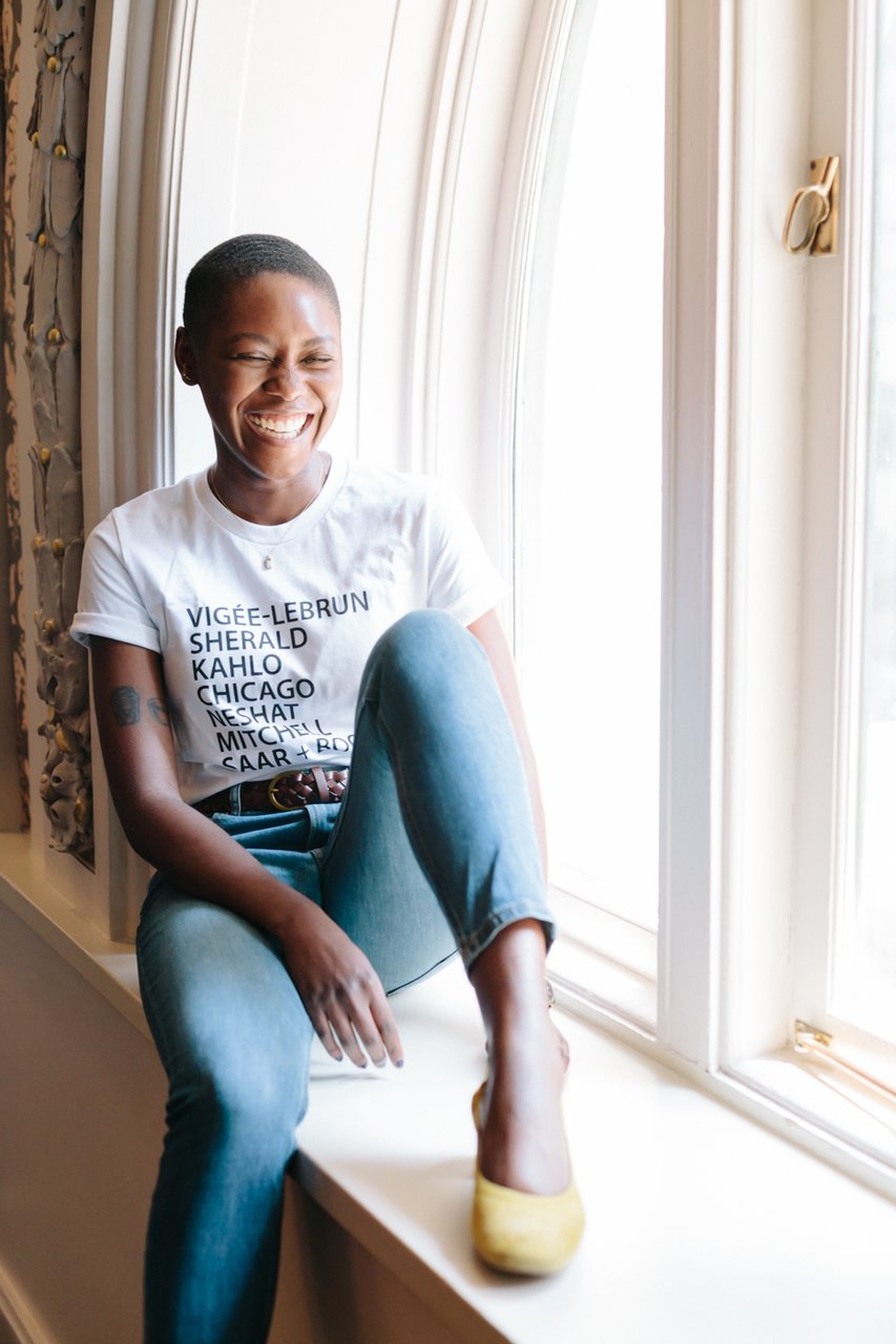 A dark-skinned woman with short cropped hair wears a t-shirt with a list of names of women artists and sits in a large window's sill. She is smiling/laughing and her eyes are closed.