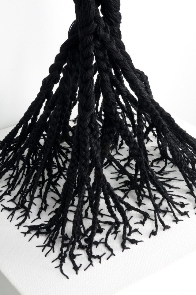 A detail image of a sculpture made from black cotton thread that has been woven into a white canvas shelf in a branch-like formation, and braided upwards. The braids are intertwined at the top.