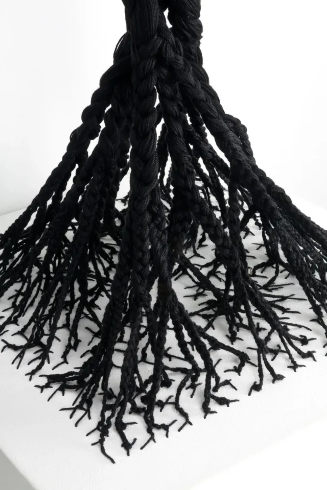 A detail image of a sculpture made from black cotton thread that has been woven into a white canvas shelf in a branch-like formation, and braided upwards. The braids are intertwined at the top.