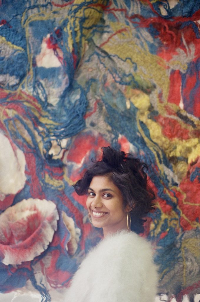 A medium-skinned woman with short wavy black hair stands smiling in front of a large textile work. She wears a white furry sweater and gold hoop earrings.