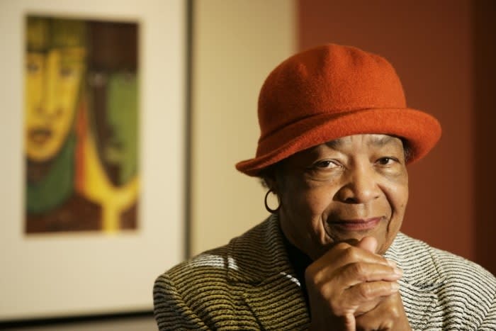 A portrait of a dark-skinned woman who wears a red felt hat, a blazer, and medium-sized hoop earrings. She stares smiling at the camera with both of her hands resting in a fist below her chin. Behind her is an abstract painting, just out of focus.