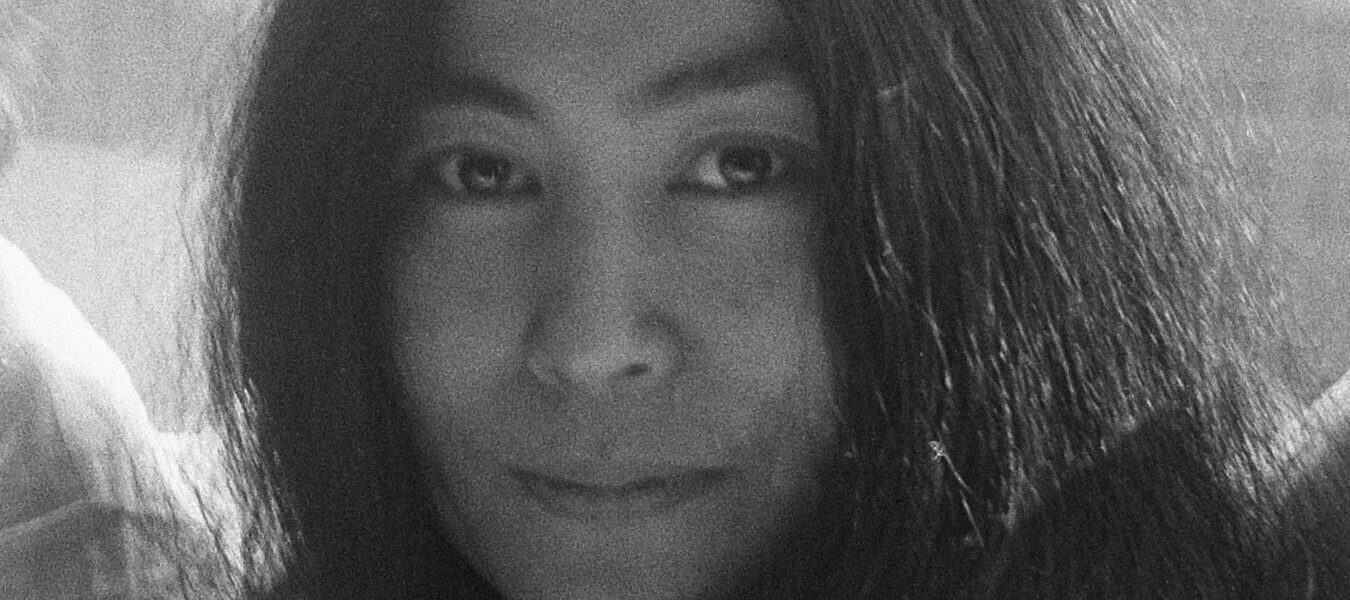 A black-and-white photograph of Yoko Ono in 1969, she wears her black hair long and sits in front of a window. The photo is cropped to her head and she smiles ever so slightly at the camera.