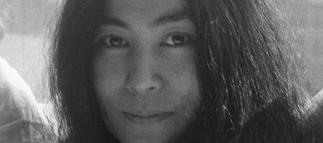 A black-and-white photograph of Yoko Ono in 1969, she wears her black hair long and sits in front of a window. The photo is cropped to her head and she smiles ever so slightly at the camera.