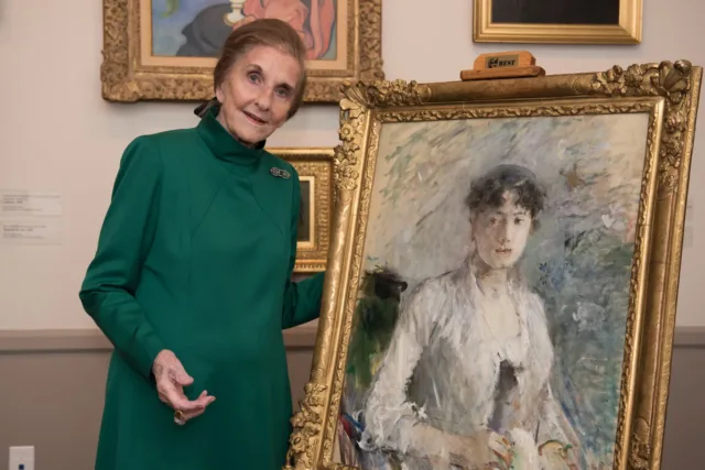 An older light-skinned woman wearing a green dress with a high neck poses next to an impressionist portrait painting of a woman with brown hair. The woman smiles and rests her hand behind the painting’s ornate gold frame.