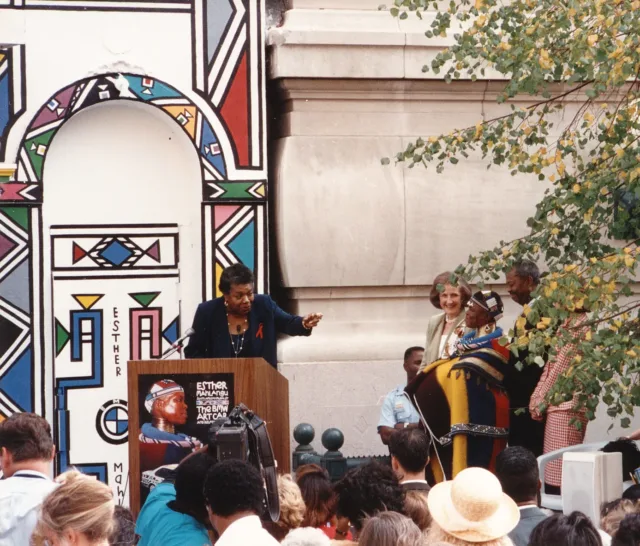 A dark-skinned woman stands at a podium which is set up outside of a large building and in front of a large crowd of people. The woman points to another dark-skinned woman to the right, who is dressed in colorful, traditional African dress and jewelry. She smiles warmly next to a light-skinned woman with shoulder-length brown hair and a dark-skinned man. Behind the podium is a large geometric mural.