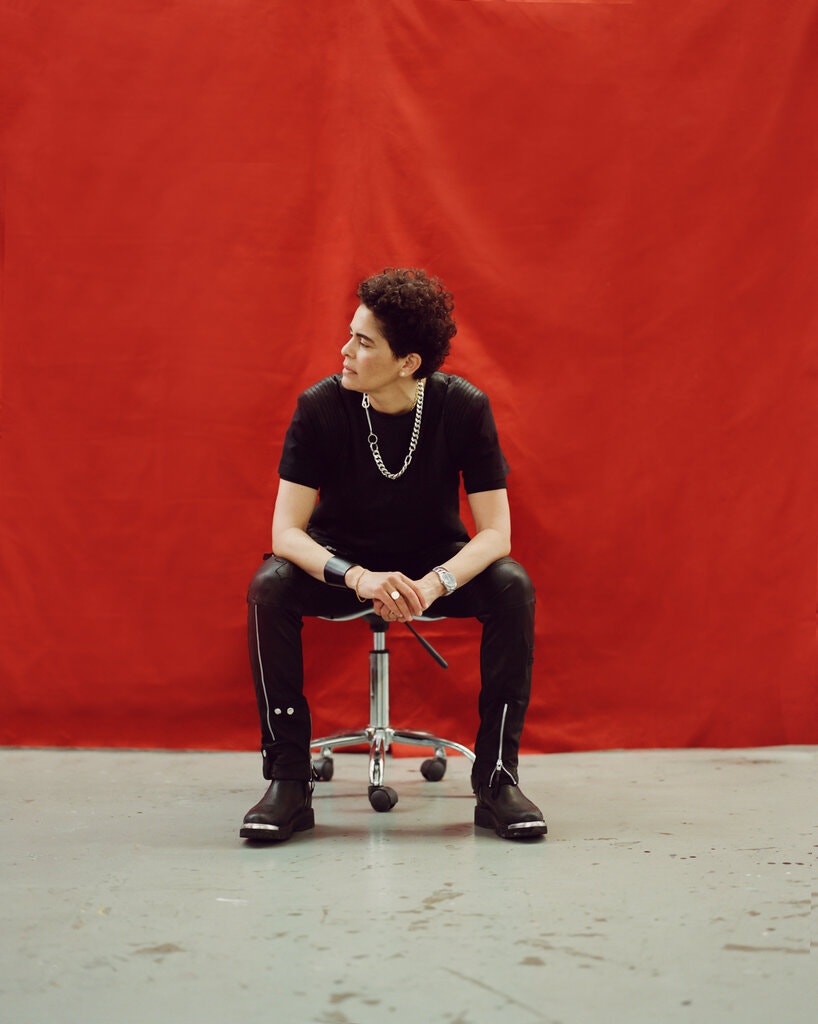 A light-skinned woman wearing a black t-shirt, black pants, and black boots, sits on a roller stool in front of a large red backdrop. Her curly hair is cut short and she looks off to the left, unsmiling.