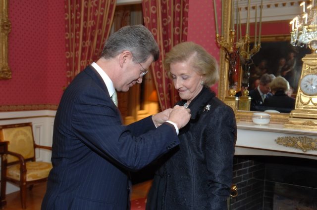 In an ornate room featuring red and gold patterned wallpaper and curtains, a light-skinned man fastens a pin to the lapel of the jacket of a light-skinned older woman. An ornate mirror and candelabra sit above a fireplace to their right.