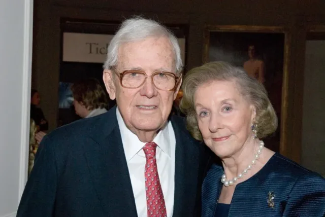 A light-skinned elderly couple smile slightly for the camera, which captures them from the chest up. They are dressed in formal evening attire, the woman wearing large pearls, a broach, and gemstone earrings. The man wears a red patterned tie and glasses.