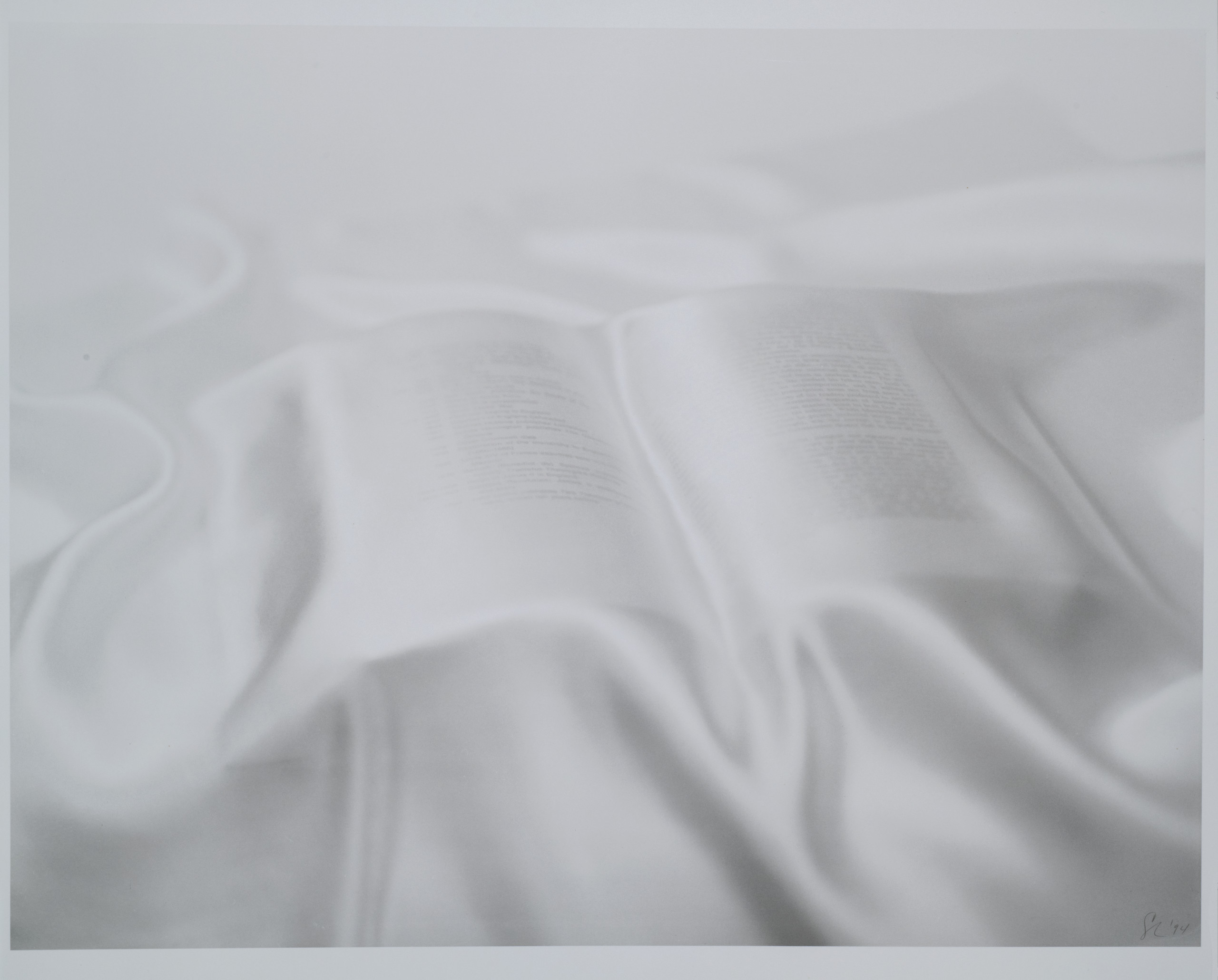 A pure white satin or silk sheet is draped over an open book. Lines of black text is visible through the sheet.