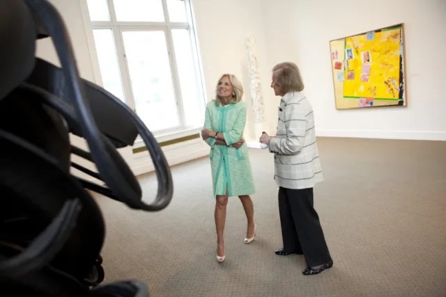 In a large, light-filled gallery, two women look on at a wall sculpture that is made of black shredded tires. The woman on the left wears a turquoise blue skirt and jacket set with white open-toed heels. She smiles looking on at the artwork. The women on the right is older and dressed in black pants and a white and grey striped over coat. The appear mid-conversation.