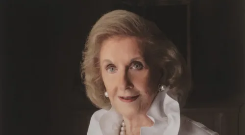 Professional portrait of a light-skinned, older woman with a blonde bob. She wears an elegant white ruffled blouse and pearl jewelry.