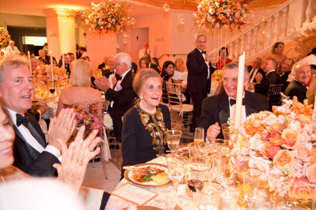 An older light-skinned woman is celebrated during a gala dinner. Many guests sit in the seats and turn towards her clapping. She smiles happily. Large, ornate bouquets decorate the hall and are the centerpieces of the tables.