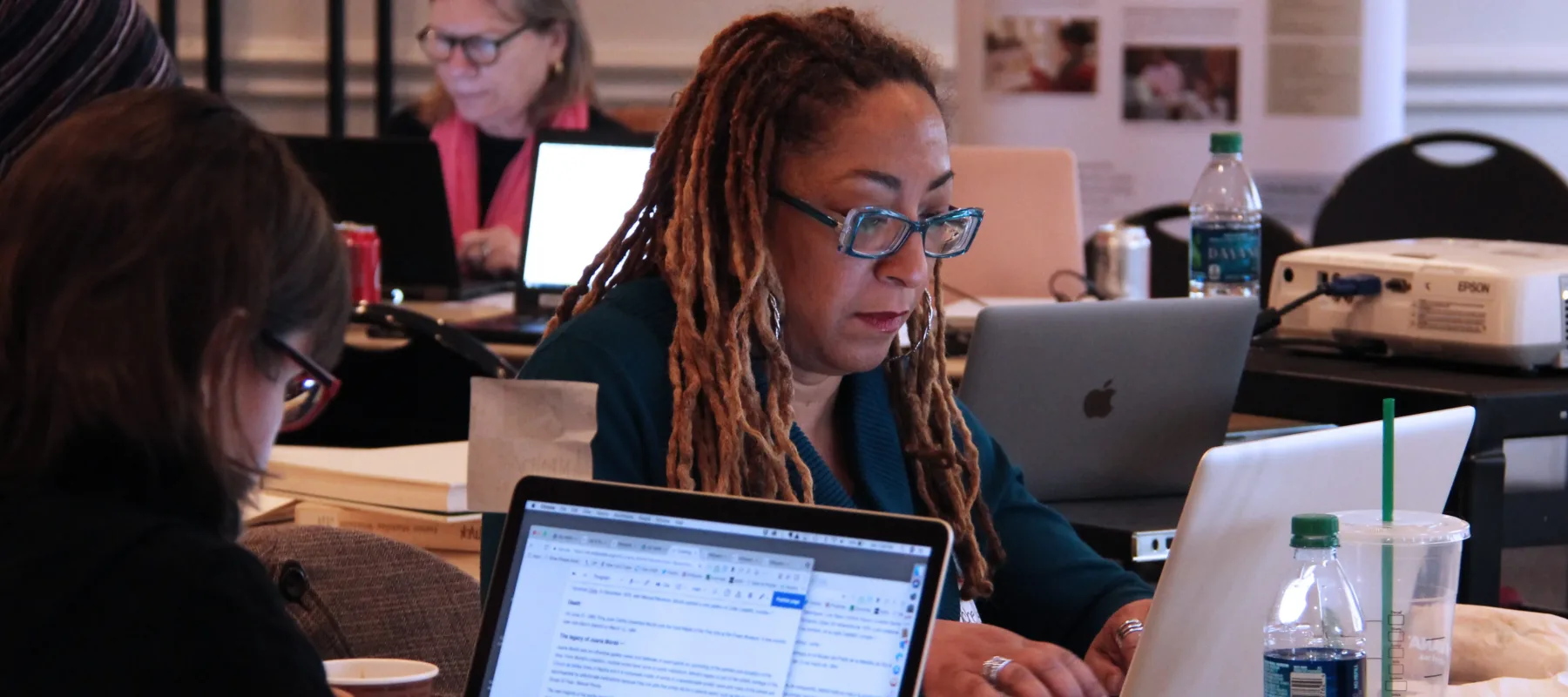 A woman with ombré dreadlocks sits at a table working on her laptop. Two other women are doing the same thing at different tables directly behind and in front of the woman.