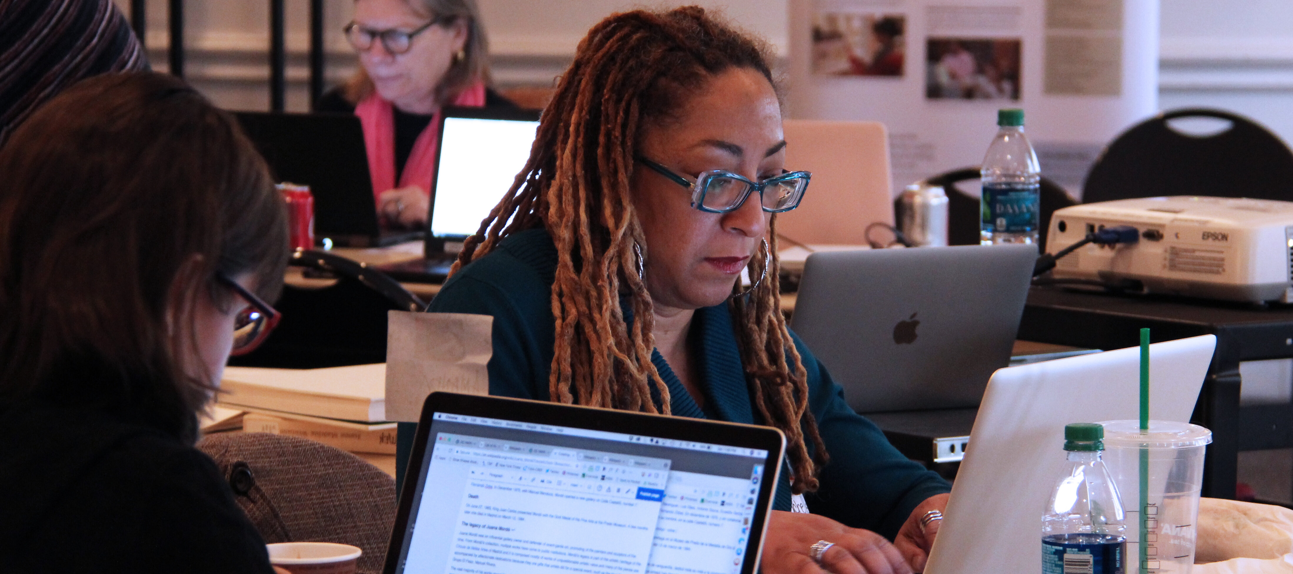 A woman with ombré dreadlocks sits at a table working on her laptop. Two other women are doing the same thing at different tables directly behind and in front of the woman.