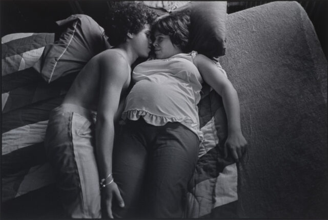 A black and white photograph of two teenagers lying next to each other on a striped blanket. The girl is pregnant and wears a light colored tank top and dark pants. She has short, dark, straight hair and lies on her back. The boy is shirtless and wears light colored pants. He has short, dark, curly hair. He lies on his side next to the girl with his hand on her leg. Their faces are nearly touching as they look into each other’s eyes.