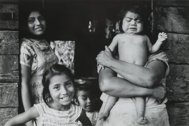 A black and white photograph of women and children standing in a wooden doorway. A figure whose head and face are obscured holds a baby, who is crying. Next to her stands a woman who leans against the doorframe, smiling. In the lower part of the frame are two young girls. The one in the foreground smiles broadly, her dark hair pulled back from her face.