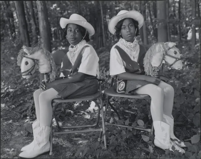 A black-and-white photograph of two dark-skinned young twins sitting in folding chairs against a woodsy background, facing the camera with their bodies angled outward in opposite directions. They wear matching cowgirl outfits, complete with hats, boots, and hobby horses.