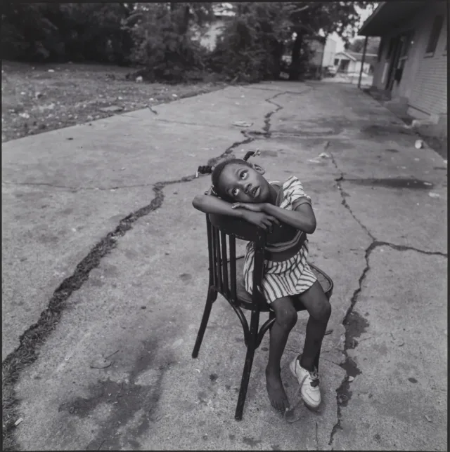 A black-and-white photograph of a dark-skinned young girl sitting sideways in a chair on a cracked street. She has braids and wears a striped dress and one sneaker. Her arms rest atop the back of the chair, and her head rests sideways on top of her arms as she looks skywards.