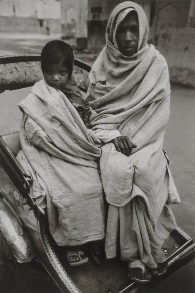 A black and white photograph of a young girl and older woman sitting side by side in a wheeled cart. The young girl has dark hair, which is pulled back, and she is wrapped in a light colored blanket. The older woman is wrapped entirely in a light colored blanket from her ankles to her head. Her hair and body are not visible. They both stare directly into the camera with serious expressions.