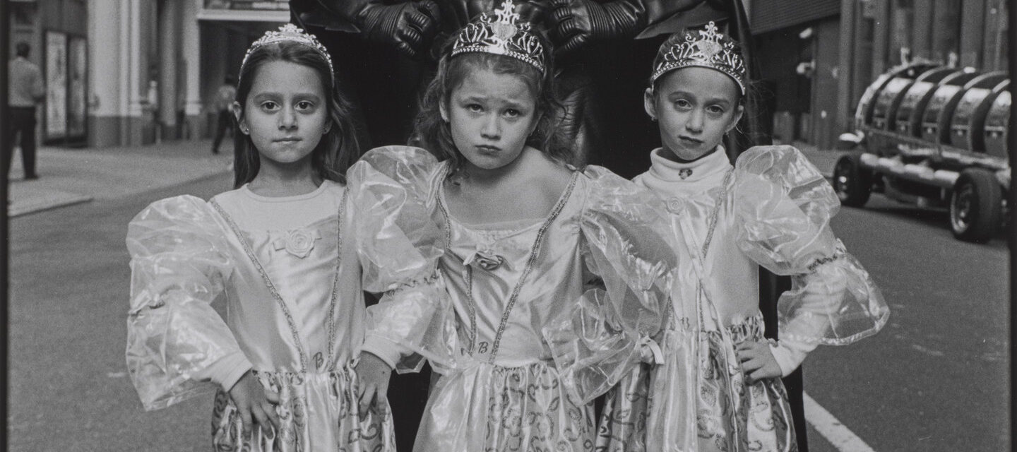 A black-and-white photograph of three light-skinned young girls in princess costumes of dresses and crowns standing in front of an adult in a full Batman costume. They all stand in the middle of a city street with defiant poses, their hands on their hips and faces serious.