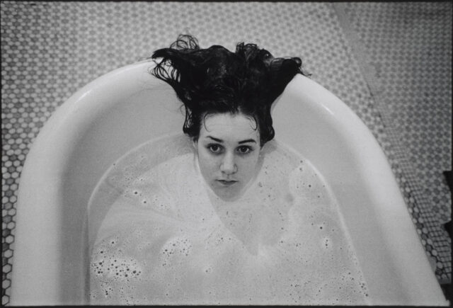 A black-and-white photograph of a light-skinned girl submerged in a white bathtub. Only her head is visible above the soap suds, and her dark hair hangs over the side of the tub. The floor beneath the tub is tiled.