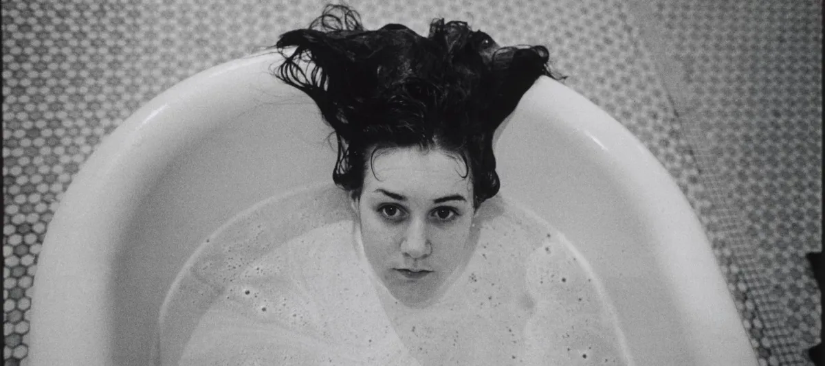 A black-and-white photograph of a light-skinned girl submerged in a white bathtub. Only her head is visible above the soap suds, and her dark hair hangs over the side of the tub. The floor beneath the tub is tiled.