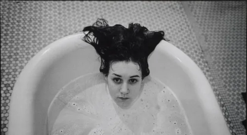 A black-and-white photograph of a light-skinned girl submerged in a white bathtub. Only her head is visible above the soap suds, and her dark hair hangs over the side of the tub. The floor beneath the tub is tiled.