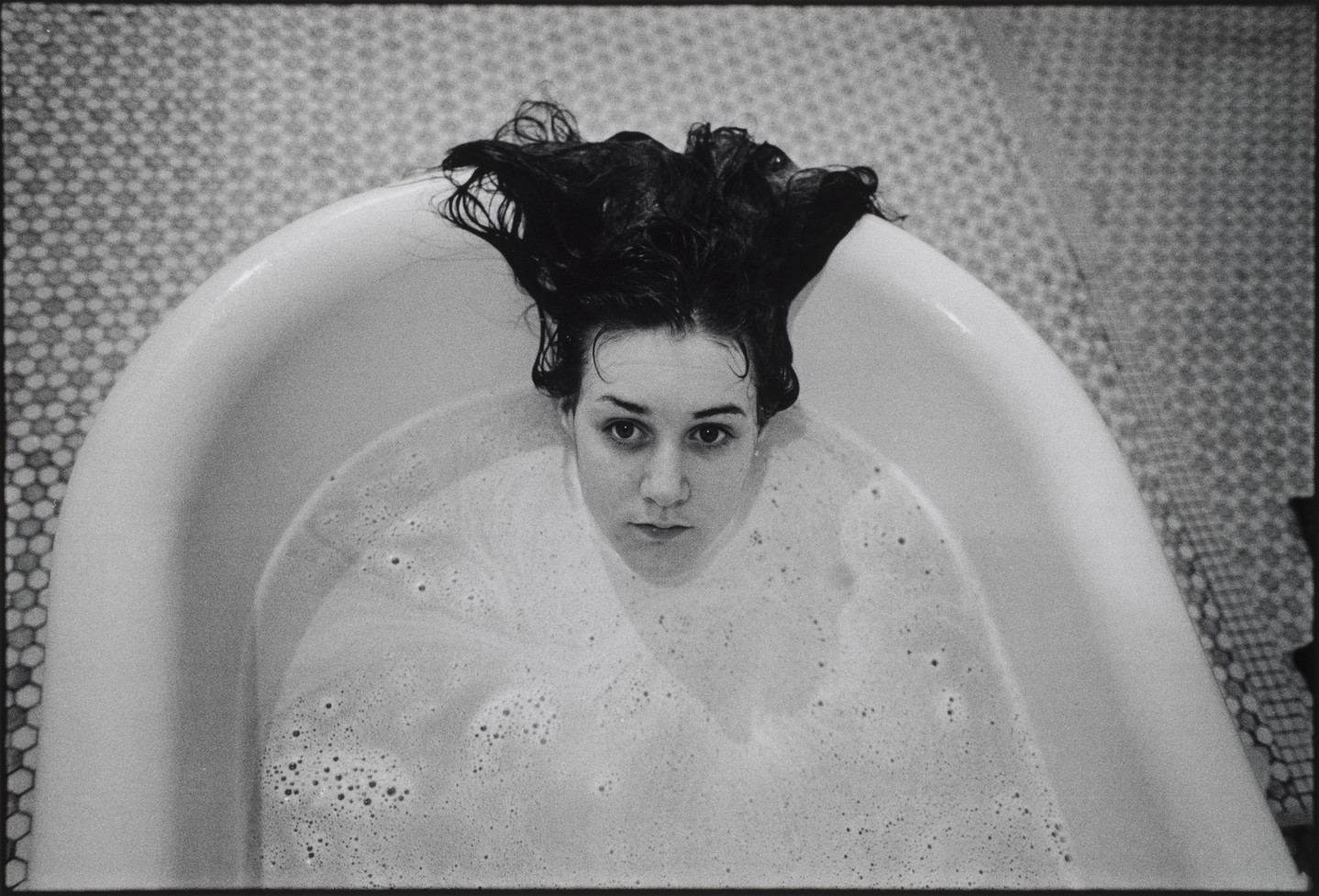 A black and white photograph of a girl submerged in a white bathtub. Only the girl’s head is visible above the soap suds, and her dark hair hangs over the side of the tub. The floor beneath the tub is tiled.