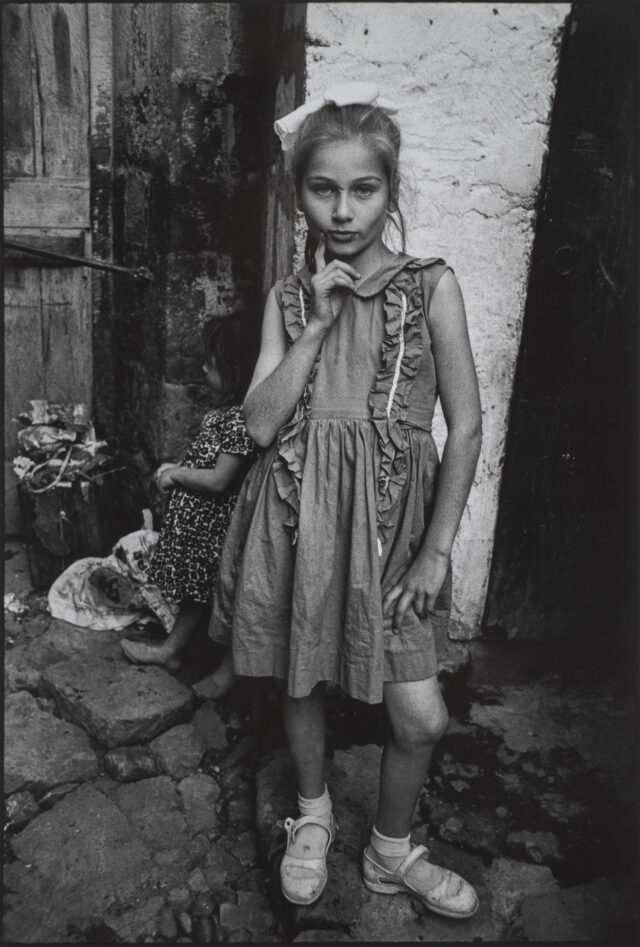 A black and white photograph of a young girl posing with her hip out and hand on her chin. She stares directly into the camera while wearing a ruffled dress, hair bow, and buckled shoes. Behind her is a stone wall that a younger child leans against.