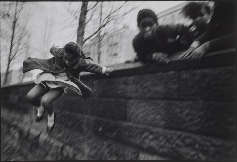 A black and white photograph of a young girl wearing a dress and coat jumping over a brick wall. Three other figures lean over the wall in the upper right corner of the composition. The jumping girl is the only figure who is in focus.