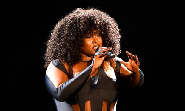 A dark-skinned woman with a large, curly, relaxed afro stands on stage in a black bodysuit with patterned cutouts. She holds a microphone up to her lips and gestures with her free hand.