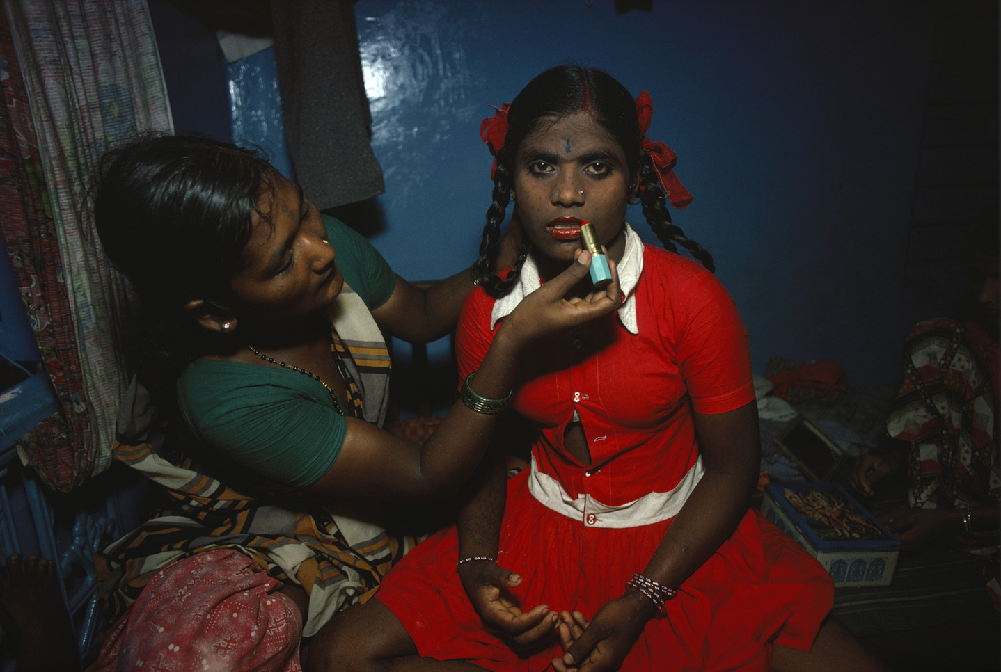A color photograph of a teenage girl sitting down next to another woman, who applies lipstick to the girl’s lips. The teenage girl has dark skin and dark hair, which is styled into braided pigtails. She wears a bright red dress with a white collar. One button in the middle of her dress is open, revealing a small amount of her abdomen. She stares directly into the camera with a blank expression.
