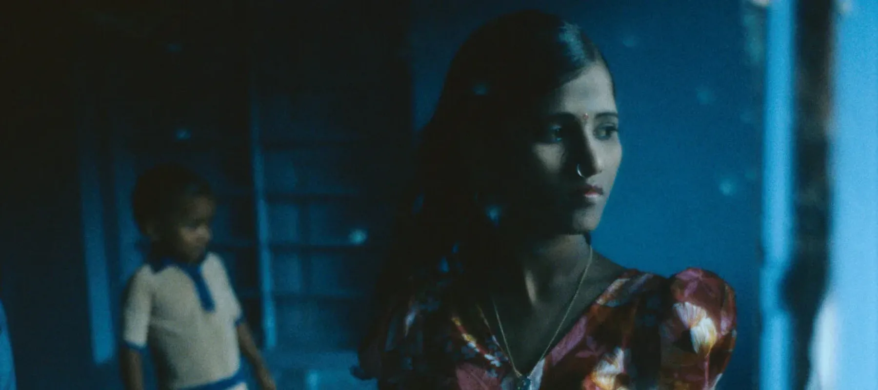 A dresser mirror reflecting a medium dark-skinned woman with long, dark hair in a vibrant red, yellow, and white dress. She faces slightly right, half of her in shadow, and wears a bindi and nose ring. Behind her is a medium-dark skinned child and they are in a very blue room.