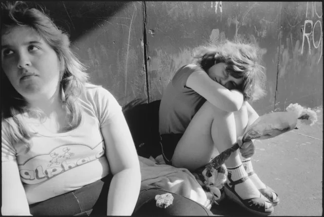 A black-and-white photograph of two light-skinned girls sitting outside against a wall. The girl on the left stares out of the frame and wears a t-shirt with 'alpha' on it. The girl to the right has her legs folded to her chest, head and arm on her knees, and holds a small flower bouquet.