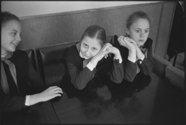 A black-and-white photograph of three light-skinned schoolgirls in dark, long-sleeved, matching uniforms. The girl on the right stares off, hands clasped by her cheek; the girl in the middle stares at the viewer with her head on her hands; and the girl on the left is laughing.