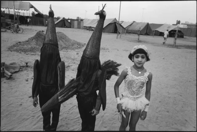 A black-and-white photograph of a medium-skinned girl in a frilly acrobat costume standing next to two children in identical, full-body peacock costumes. One peacock-costumed child holds a bundle of long peacock feathers, and they all stand on sand with a row of tents behind them.