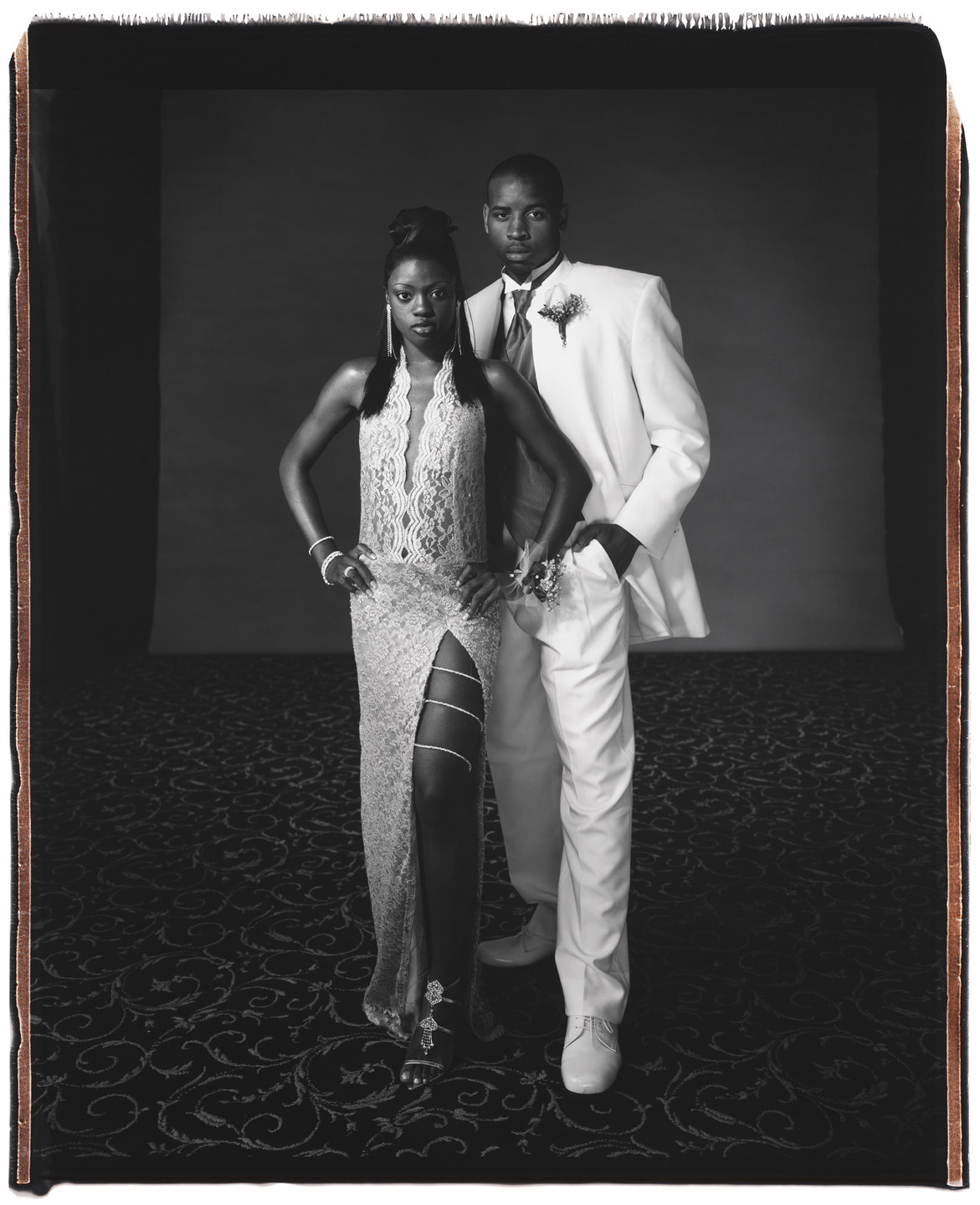 A black and white photograph of a teenage couple in formal clothing. A teen girl with dark skin and straight, dark hair stands slightly in front of a teen boy with dark skin and shortly cropped dark hair. She wears a long, light colored lace dress with a slit on the leg, and a jeweled chain wrapped around her thigh. He wears a white suit with a dark tie.