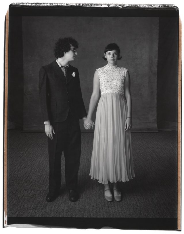 A black and white photograph of a teenage couple in formal clothing standing side by side, holding hands. The boy has light skin and short, dark, curly hair. He wears a dark colored suit with a white boutonniere. He turns his head to look at the girl, who also has light skin and short, dark, straight hair and wears a light colored, ankle length dress.