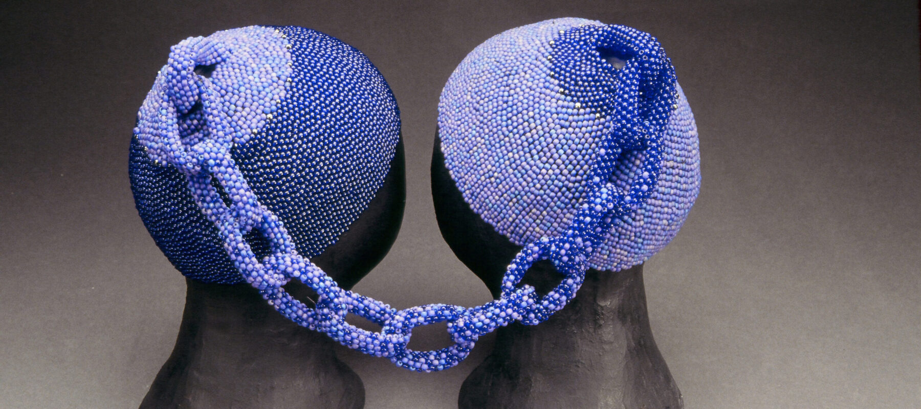 Two head caps made of small, blue glass beads rest on two black mannequin heads. The two caps are connected at the tops by a beaded chain. The left cap is made of darker blue beads and the right cap is made of lighter blue beads. The chain combines both shades.