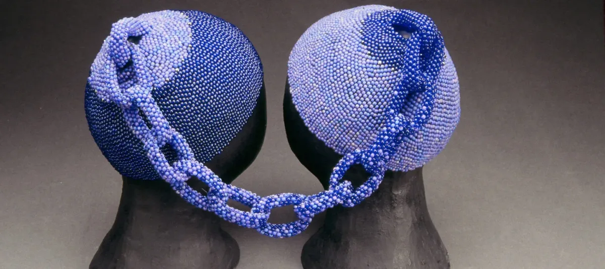 Two head caps made of small, blue glass beads rest on two black mannequin heads. The two caps are connected at the tops by a beaded chain. The left cap is made of darker blue beads and the right cap is made of lighter blue beads. The chain combines both shades.