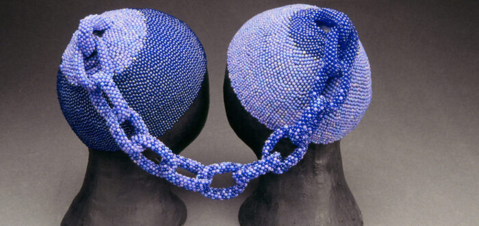 Two head caps made of small, blue glass beads rest on two black mannequin heads. The two caps are connected at the tops by a beaded chain. The left cap is made of darker blue beads and the right cap is made of lighter blue beads. The chain combines both shades.