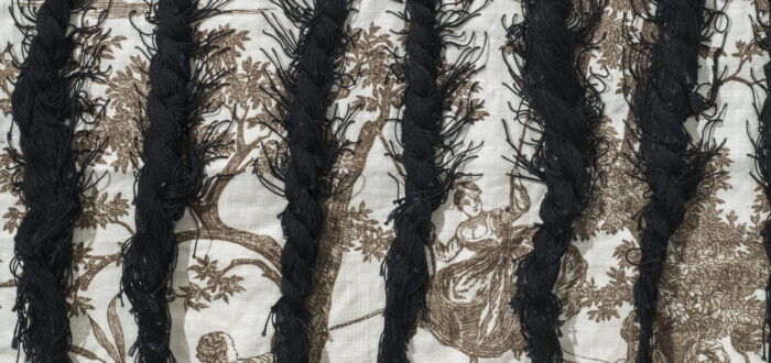 A close up photograph of braided black hair woven into cloth featuring colonial narrative scenes.