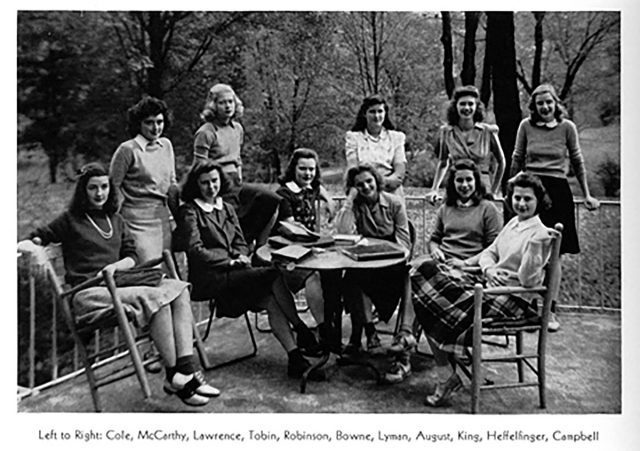 Eleven young light-skinned women pose on a patio for a group portrait. Some sit around a table covered with books; others stand leaning against a railing.