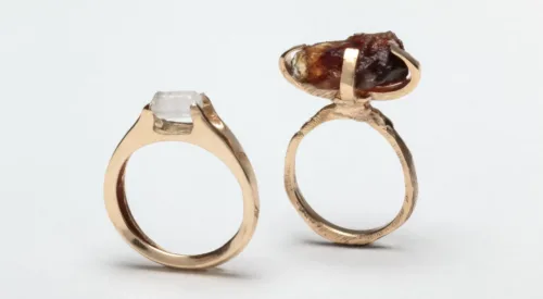 Two rings with gold bands and small gem-like forms on top. The gems are crafted from raw sugar. The left ring is perfectly circular with a small, smooth, white sugar crystal on top. The right ring is a slightly rougher circle, with a larger, lumpy, brown sugar crystal on top.