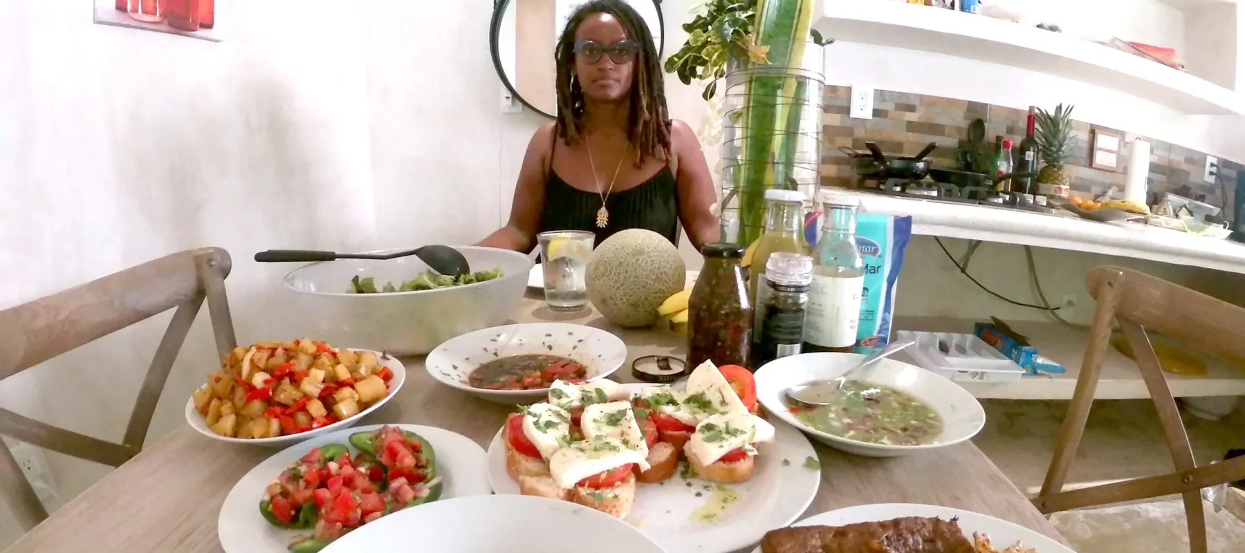 A medium-dark skinned adult woman sits at the end of a table, as if sitting across from the viewer. She has shoulder-length, dreadlocked hair, and wears glasses and a black tank top with a gold necklace. The table is set with colorful foods like avocados and bruschetta.