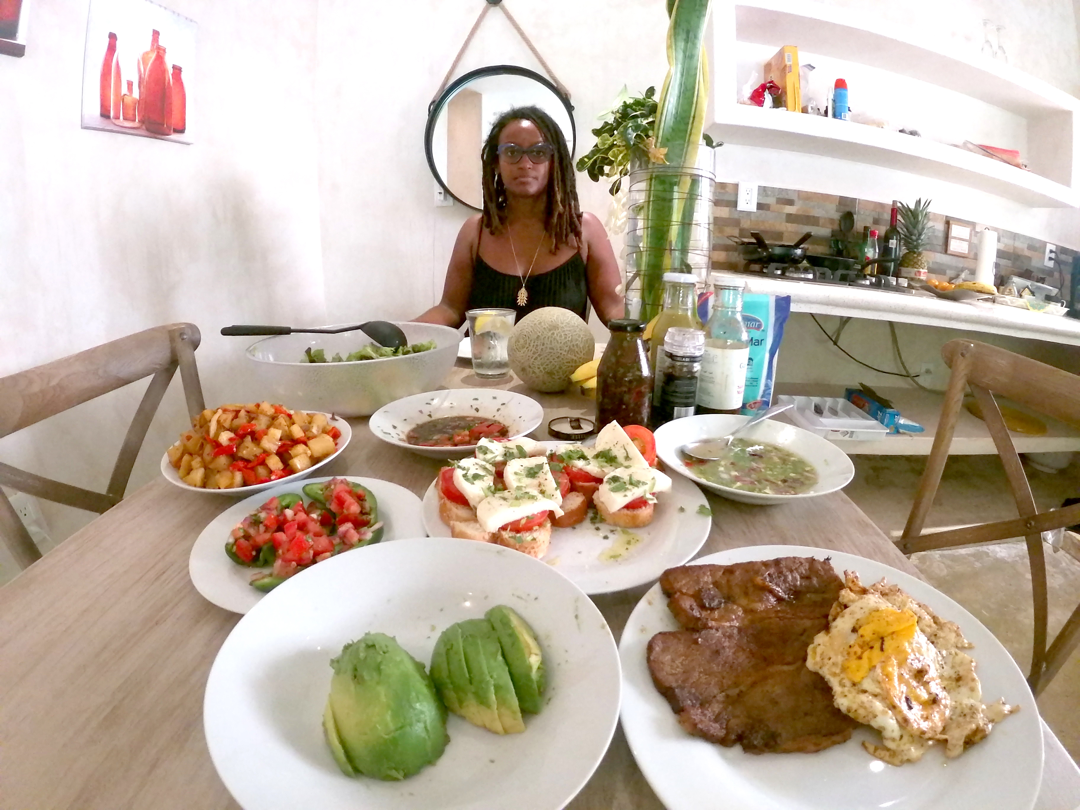A medium-dark skinned adult woman sits at the end of a table, as if sitting across from the viewer. She has shoulder-length, dreadlocked hair, and wears glasses and a black tank top with a gold necklace. The table is set with colorful foods like avocados and bruschetta.