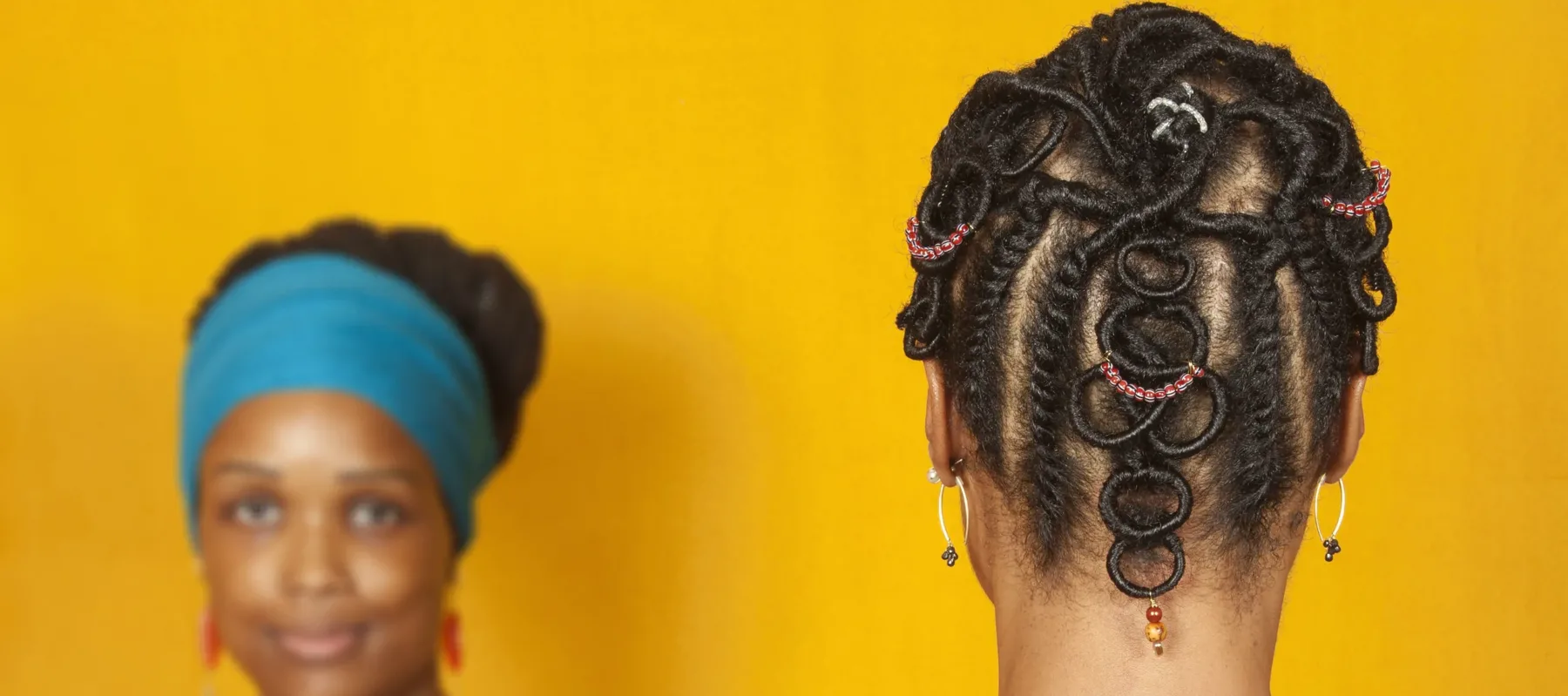 Two medium-dark skinned adult women stand in front of a goldenrod background. The woman on the left faces the viewer and wears a teal head wrap and a red top. The woman on the right faces away from the viewer, showcasing her dark hair braided into an elaborate pattern with colorful beads.