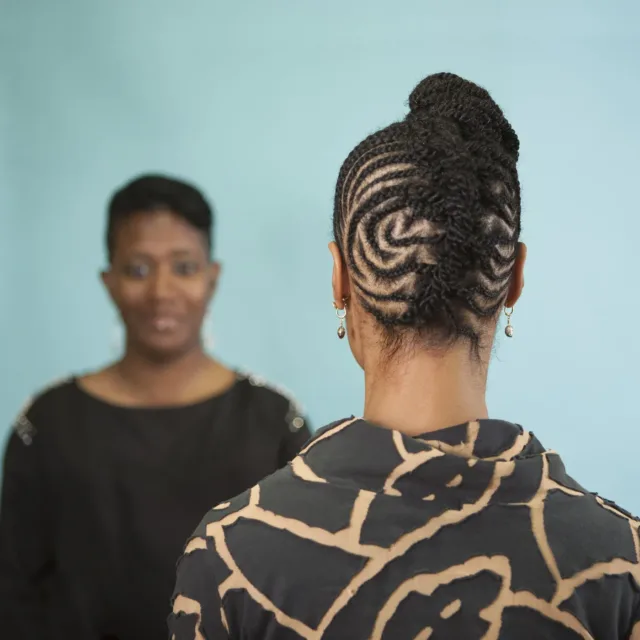 A color photograph featuring two dark skinned women in front of an aqua background. The woman on the left faces the camera. She is wearing a black shirt with gold details on the shoulders and has her brown hair pulled back. The woman on the right stands with her back to the camera wearing a black and tan patterned shirt. Her brown hair is braided in an elaborate pattern and pulled up on top of her head.