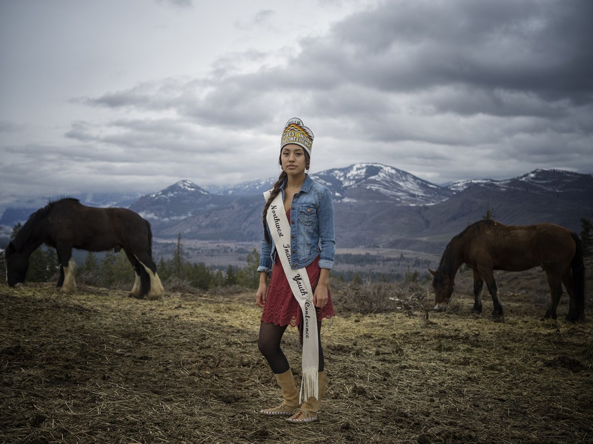 An American Indian woman stands outside under a cloudy sky, flanked by two grazing horses and with mountains spotted in snow in the background. She wears moccasin boots, vlack tights, and a deep pink dress that falls to her knees over which she wears a jean jacket. A headpiece adorns her head and a long white sash is draped over her torso.