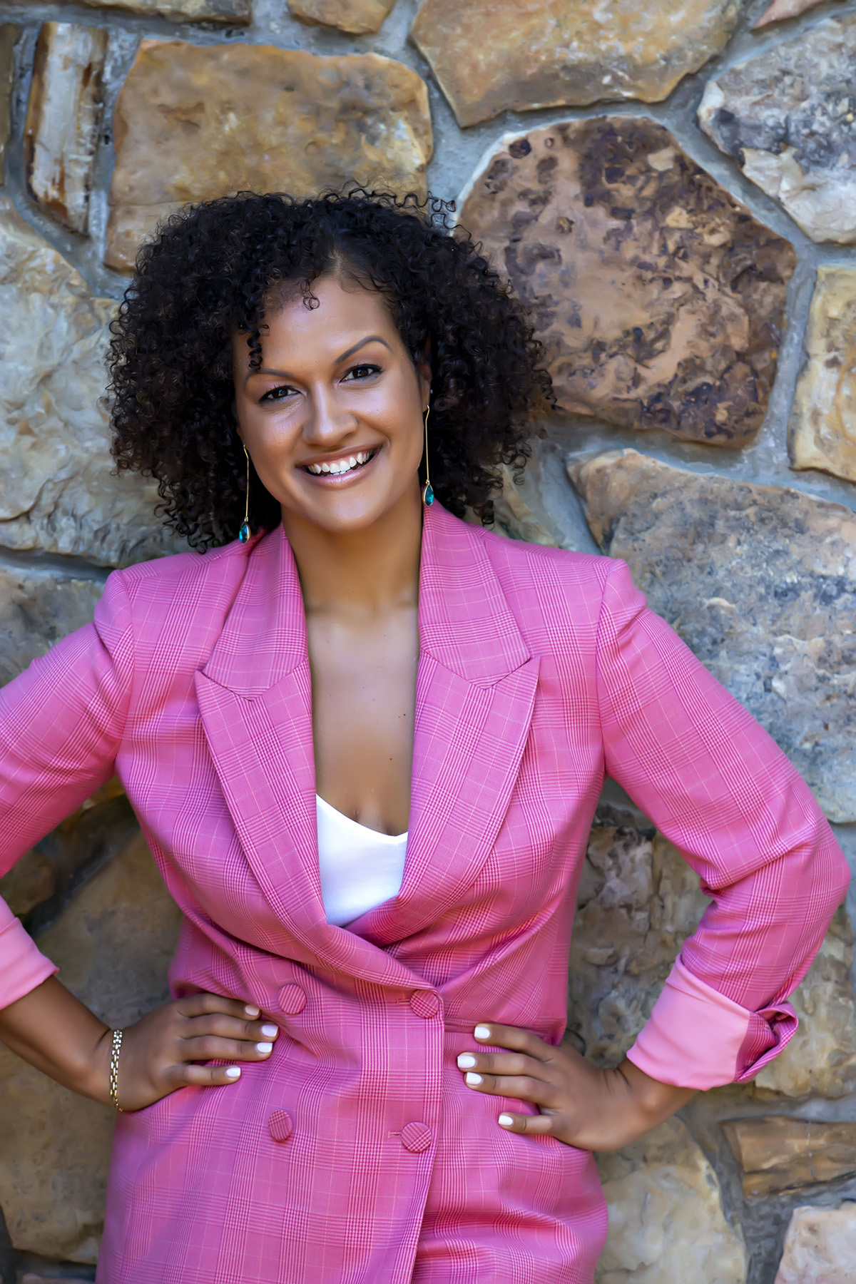 A medium-skinned woman with tight, chin-length black curls stands against a stone wall wearing a pink blazer with a low-cut neckline. She rests her hands on her hips and smiles warmly at the camera.